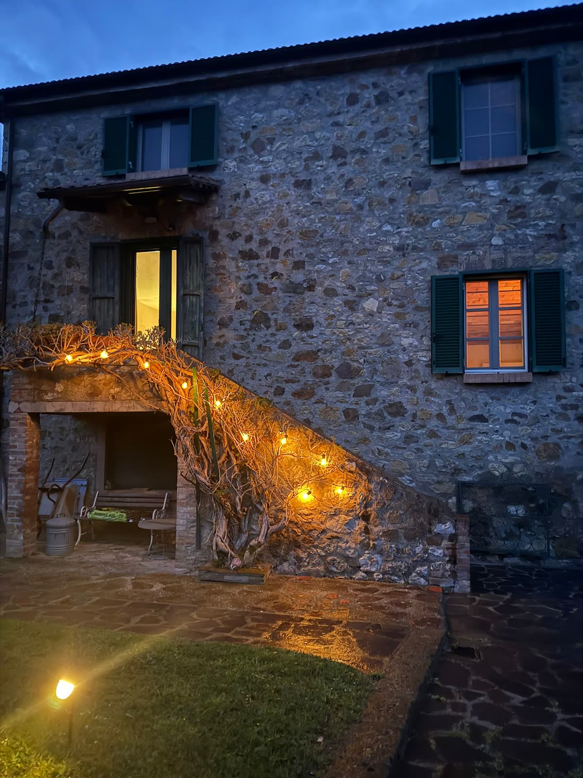 The stone house at dusk, with warm lights strung along the vine on the outside staircase and lit windows above
