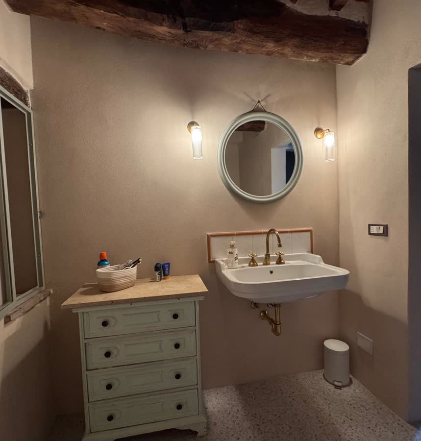 Bathroom with a round mirror, brass taps and a wall-hung basin under an old timber beam
